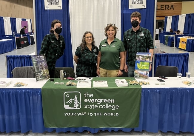 Four people stand behind a table with the words "the Evergreen State College" on a banner