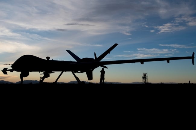 A silhouette of a military drone that looks like an airplane. The silhouette is set against a light blue sky with white clouds. A serviceperson standing beneath the rear of the drone, is also silhouetted.