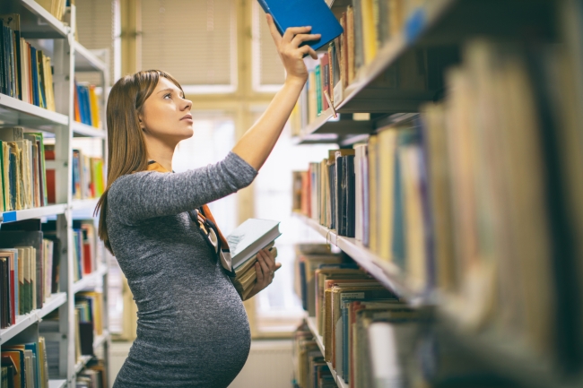 A pregnant person in a gray dress puts away a book in a library shelf