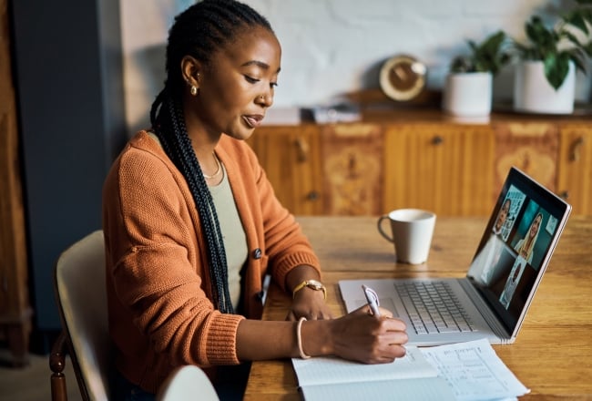 Woman sits in front of laptop and Zoom conference in home office, writing notes and listening to colleagues during online meeting.