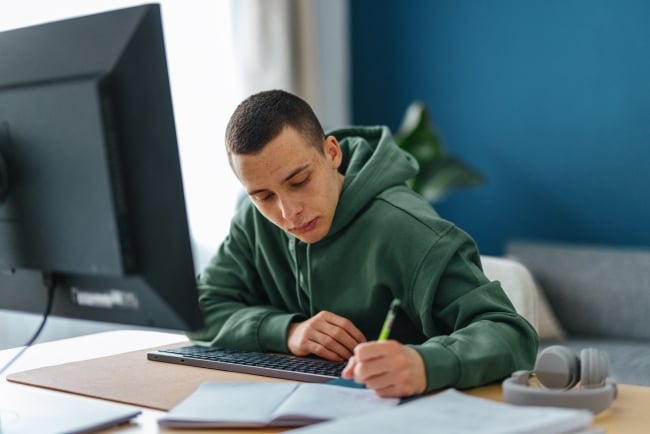 Portrait of a young male student, sitting at his desk studying for school or following an online class on his digital tablet. He is taking notes in his notebook.