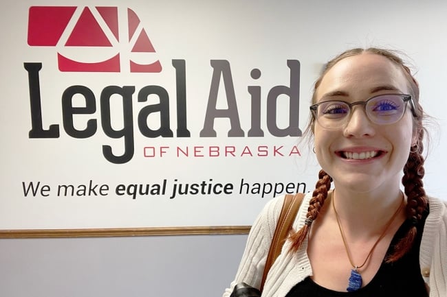 A student smiles in front of a sign reading Legal Aid of Nebraska