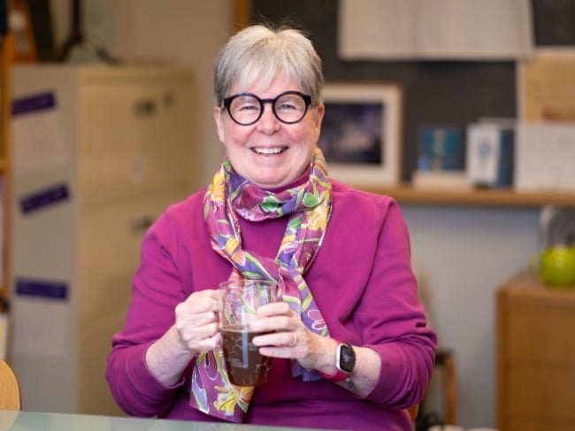 A smiling woman with gray hair and black glasses holding a large cup of tea