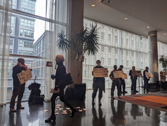 A photograph of seven demonstrators holding signs in a building as someone walks by. One sign says "Protect Pro-Palestine Activism" and another says "Stop Silencing Activism for Palestinians."