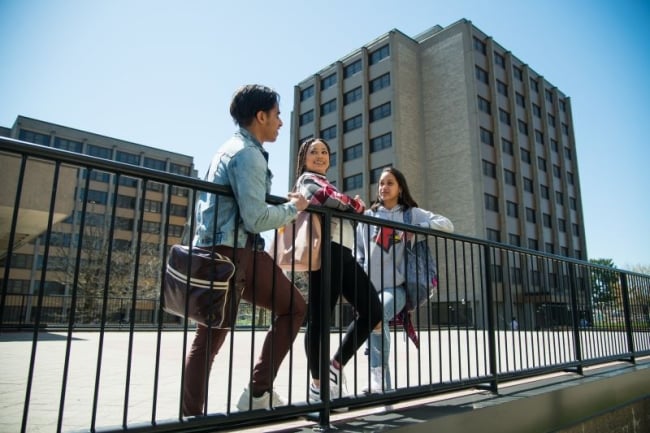 Three students lean on a railing with the SUNY Plattsburgh campus in the background.