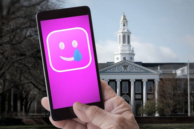 A cellphone with a pink screen and smiley face with a teardrop is held up front of a backdrop of Harvard University.