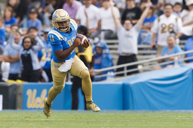 A football player in blue and gold holds a football and runs down the field