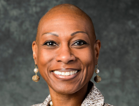 Antoinette "Bonnie" Candia-Bailey, a Black woman with a bald head, wearing a textured jacket and a statement necklace with matching earrings, smiles in a professional headshot.