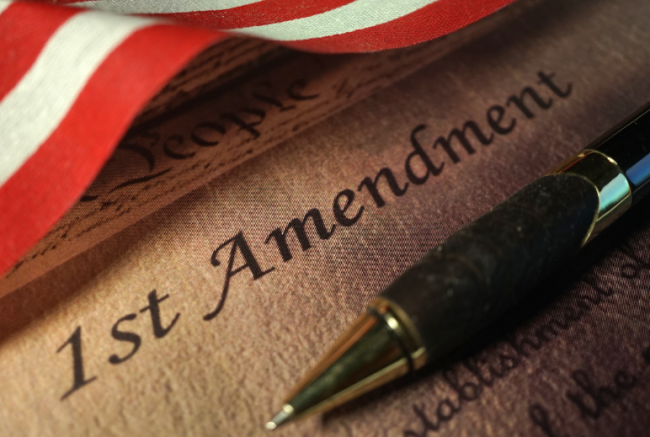A document bears the heading "First Amendment," with a pen lying atop it. The bottom of a waving American flag is visible in the upper lefthand corner of the image.