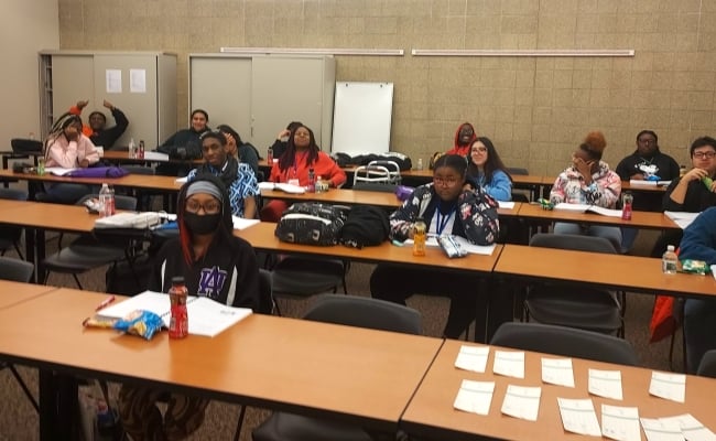 A group of young people sit at tables in a classroom doing work and smiling at the camera.