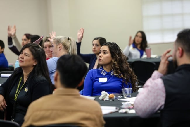 Faculty and staff sit at round tables, some raising their hands in the background. 