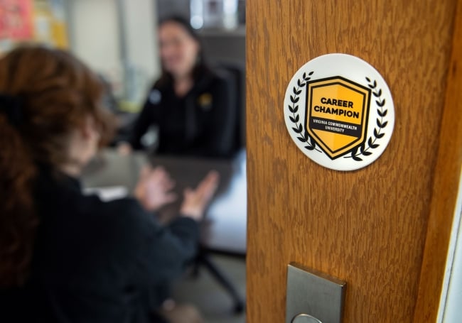 A sticker on the door to an office is in the foreground, with two people talking around a desk in the background. The sticker is white, with a yellow crest and the words "Career Champion" printed in black