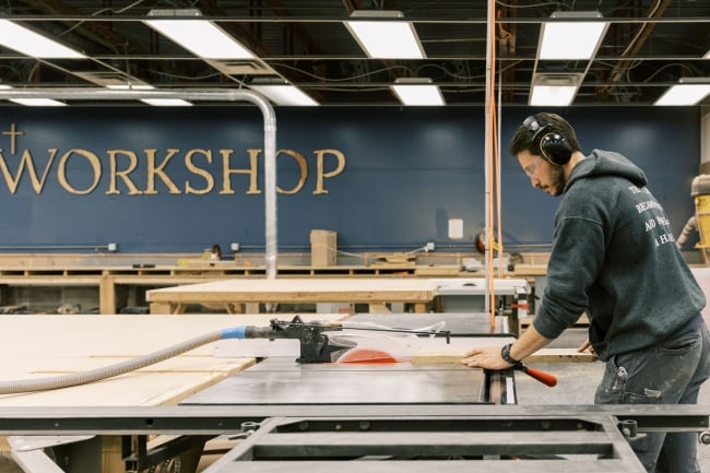 A man practices carpentry with the word "WORKSHOP" emblazoned on the wall behind him.