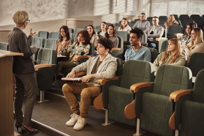 A room full of students in a lecture hall looks at a professor teaching in front of a podium. 