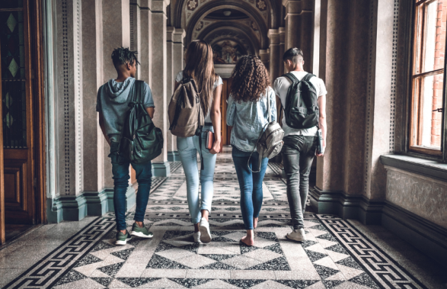 A group of four students, carrying backpacks, walk away from the camera down the hallway of a university building.