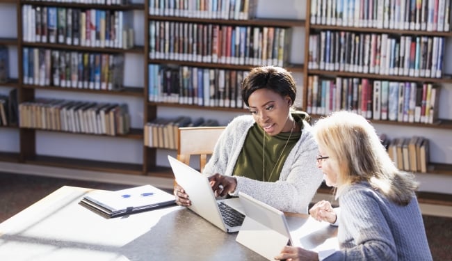 Two women working together in a library at a table. One has a laptop and the other is using a digital tablet to share information.