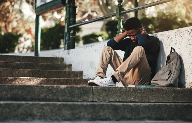 A student sits on stairs next to a backpack, looking frustrated and sad.