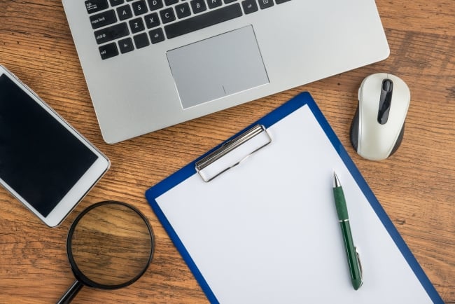 Desktop with computer, iphone, magnifying glass, and clipboard with pen and blank paper