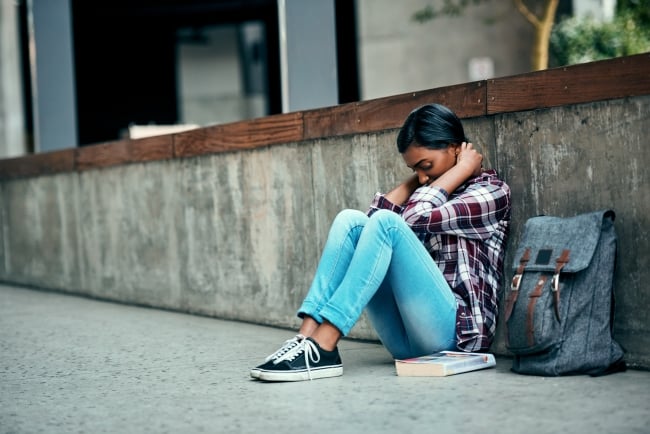 A female college student sits with her back to a wall and her head cradled in her hands, looking depressed, next to a backpack and book.