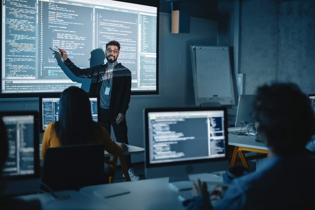 A man points toward a screen filled with code. He is presenting in a dark room filled with students working on computers.