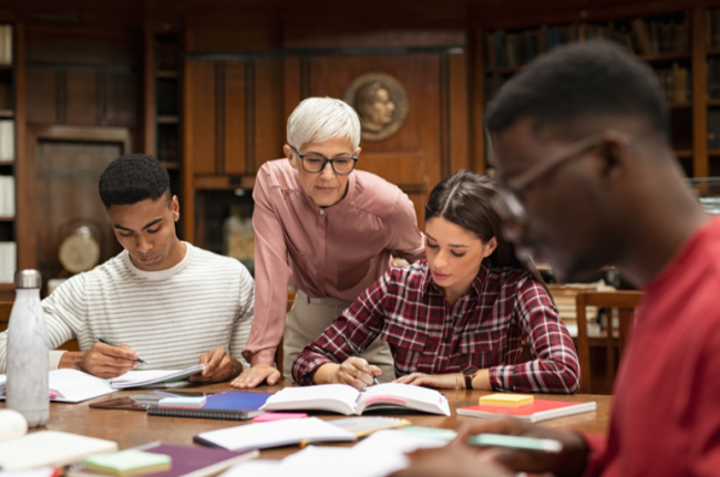 A group of three students studying at a table with the help of a professor standing behind them.