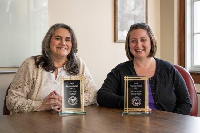 Two female professors sit behind a desk with small awards on the desk.