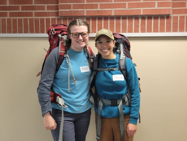 Two students wearing blue shirts and backpacks smile for a photo in front of a brick wall