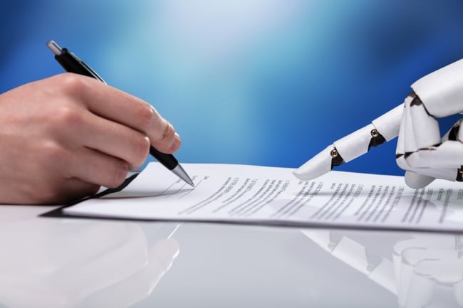 On the left side of the photo, a hand holds a pen and is writing on a white piece of paper. On the right side, a robot is pointing toward the piece of paper.