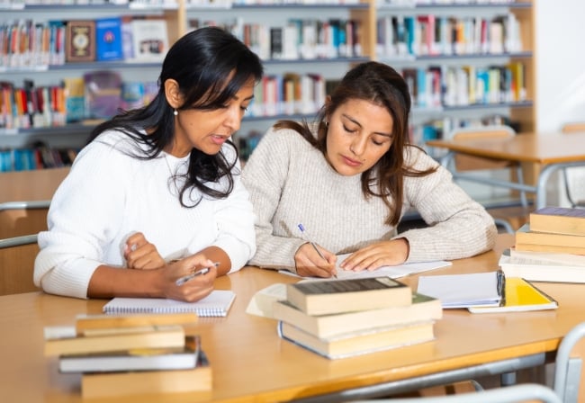 Two females students sit at a desk writing together