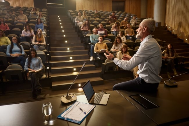 Senior professor talking in front of large group of his students during a lecture in an amphitheater.