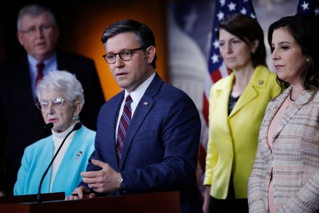 Mike Johnson, in a navy jacket, stands at a podium next to Elise Stefanik and Virginia Foxx