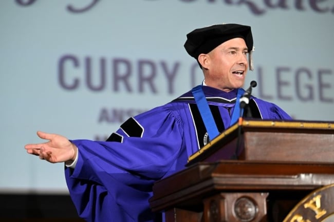 Curry College President Jay Gonzalez wears academic robes and gestures with his arms at a podium with “Curry College” projected in purple letters behind him.