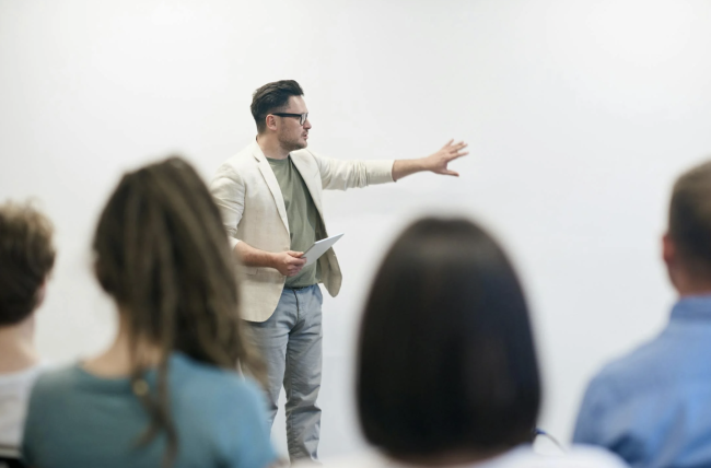 Male professor addresses a group of students before a white background.