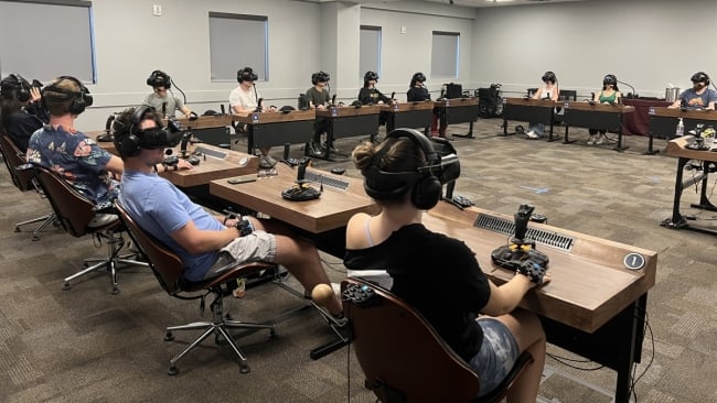 A group of students sit, all wearing virtual reality headsets. They are in a classroom and in a square formation with their desks. 