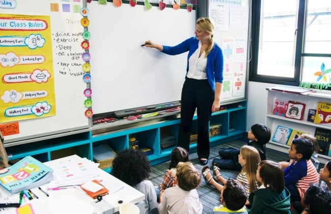 A young female teacher in a brightly decorated elementary classroom writes on a whiteboard, as the young students, sitting on the floor, pay close attention.