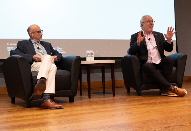 Two men sit on a stage at a conference. The man on the right is speaking and gesturing with his hands.