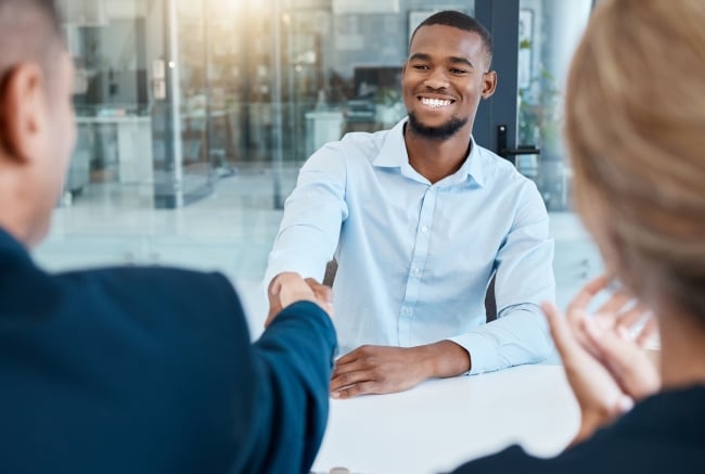 A young man shakes hands with a person across the table, smiling, in an office.