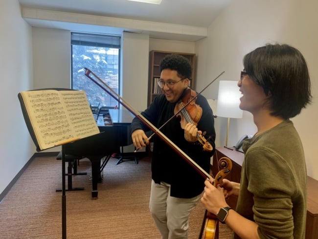 A student plays violin while smiling and looking at sheet music. 