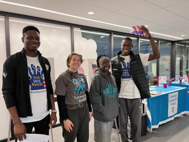 Four students stand in front of a table with a table banner that reads “Cuyahoga Community College.” Two wear shirts with the slogan “Your voice matters. #voiceyourvote.”