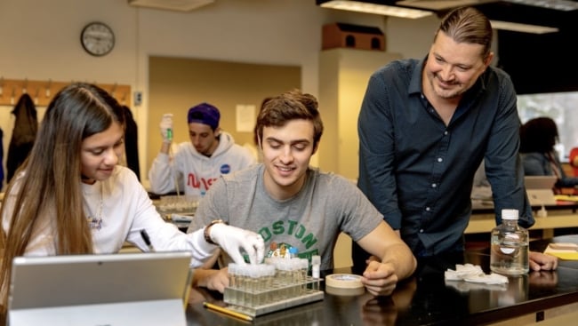 Mark Sarvary stands next to two seated students working with test tubes in a lab course setting, smiling. 