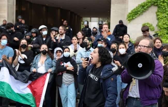 A group of twenty or so protesters gathered in a doorway. In the left bottom corner of the frame some protesters hold an unfurled Palestinian flag. One speaker in front carries a bullhorn, while another appears to be shouting into a microphone. Police officers wearing helmets and face shields are standing in the background.