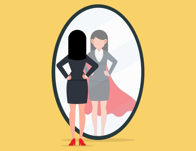 Professional woman wearing a suit stands in front of mirror examining her reflection, in which she is also wearing a cape like an action hero