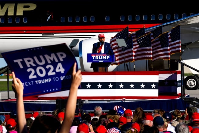 Former president Trump stands in front of a plane at a podium. A Trump 2024 sign is in the foreground.
