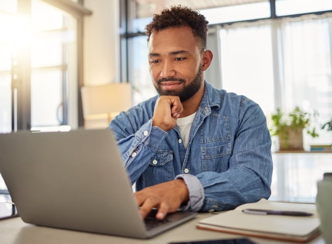 An adult man works at a laptop with a notebook and pen beside him.