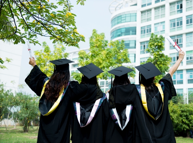 Rear view of four female graduates wearing graduation caps, gowns and hoods on campus.