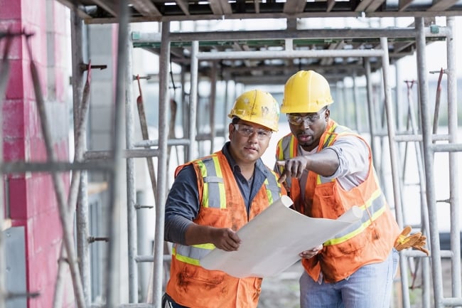 Two men in hard hats and high-visibility vests lean over a blueprint, one pointing.