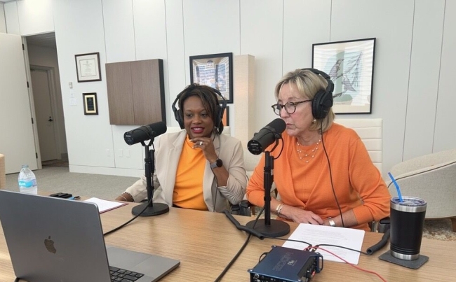 Amber Williams and Donde Plowman sit next to each other, wearing University of Tennessee orange, with microphones at the table in preparation for a podcast interview.
