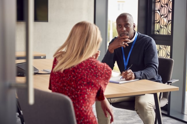 A female student meets one-on-one with a male professor. They are seated across the table, and the student can only be seen from behind; the professor is listening intently to what the student is saying and has his hand poised over a notebook.
