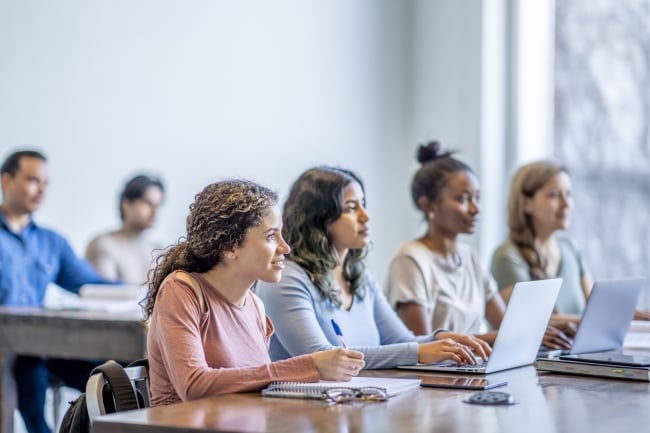 A class of University students sit together at long desks, each with notebooks and laptops out in front of them as they take notes during the lecture. They are all dressed casually and are listening attentively as they look to the front of the class and soak in all the information.