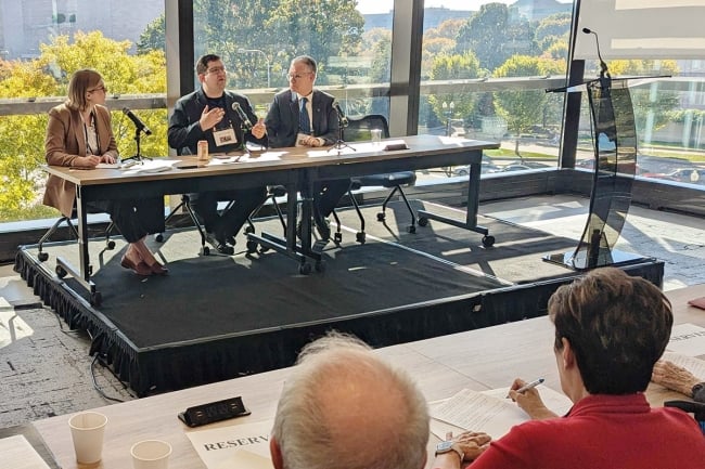 A photograph of three people on a panel sitting at a table, with large glass windows behind them.